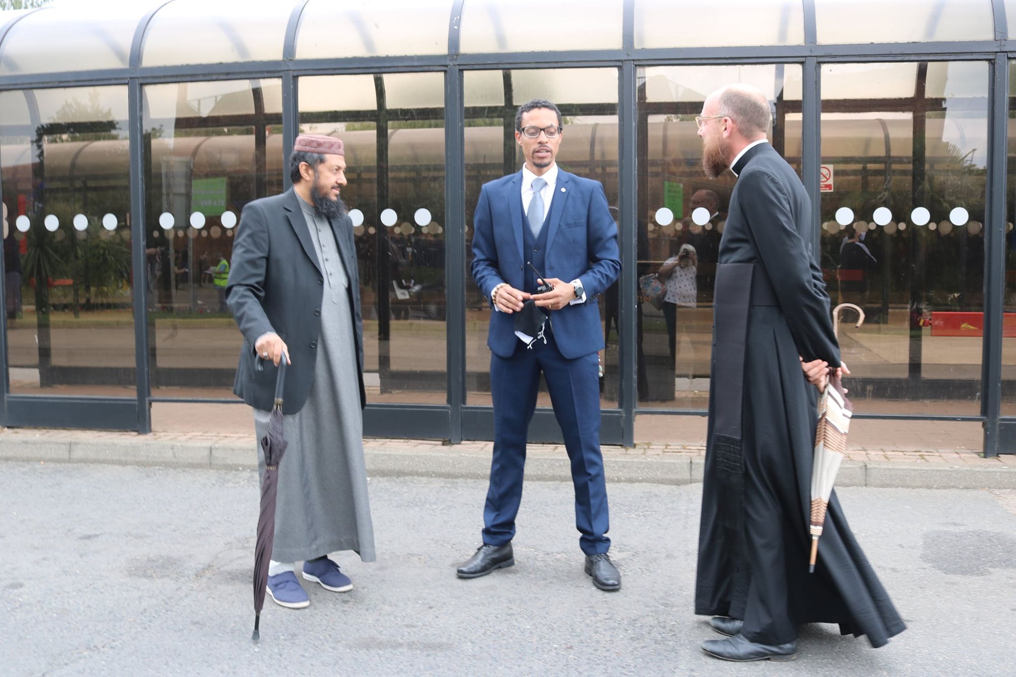JB Webb-Benjamin in a navy suit talking with an imam and a Christian priest outdoors by a bus shelter.