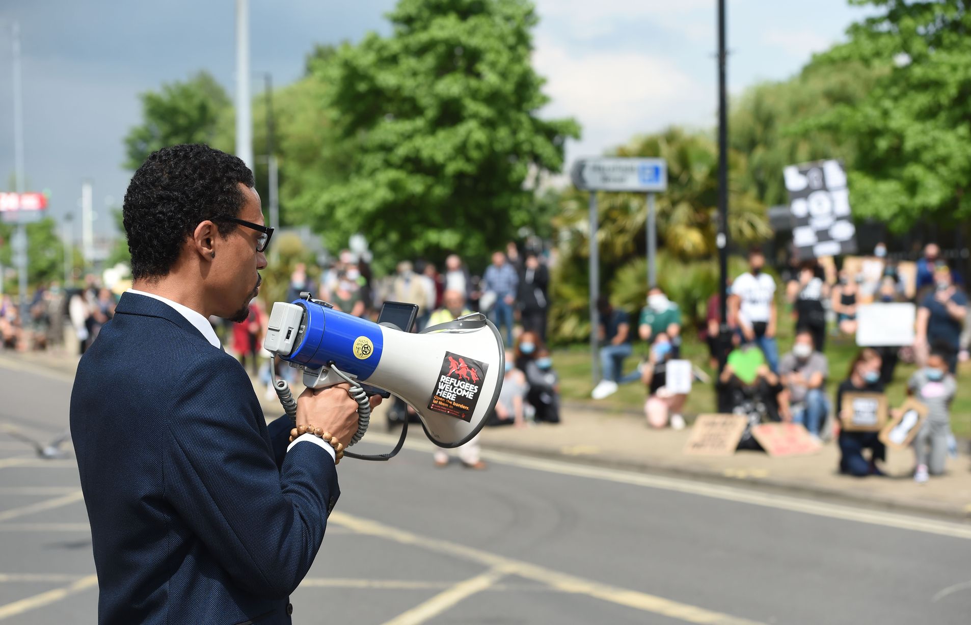 JB Webb-Benjamin in a navy suit speaking through a megaphone to a distanced, masked crowd at an outdoor rally; the megaphone carries a 'Refugees Welcome Here' sticker.