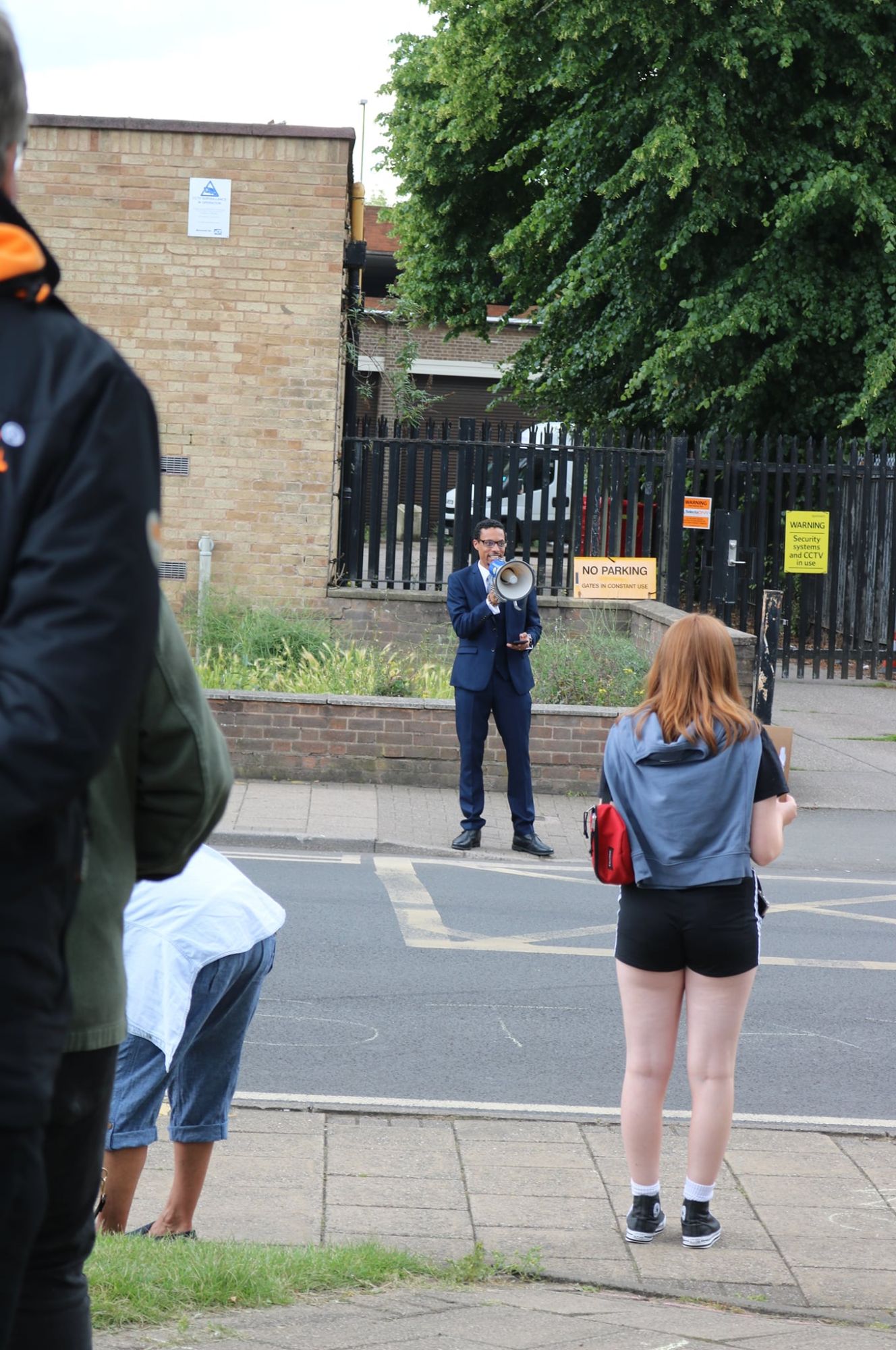JB Webb-Benjamin addressing a small socially-distanced crowd with a megaphone across a street, onlookers in the foreground.
