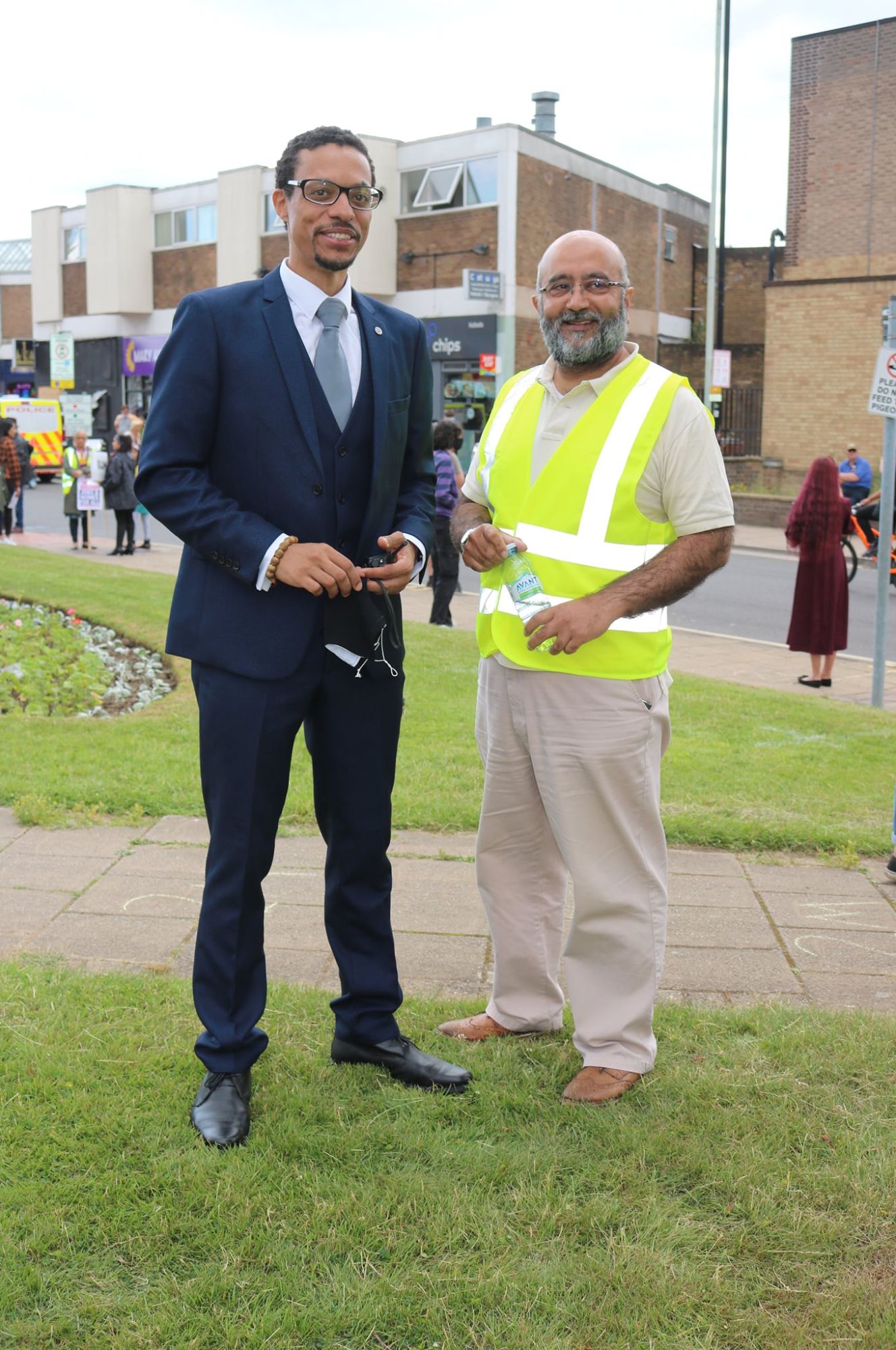 JB Webb-Benjamin in a navy three-piece suit standing on grass beside a smiling volunteer in a hi-vis vest at a community event.