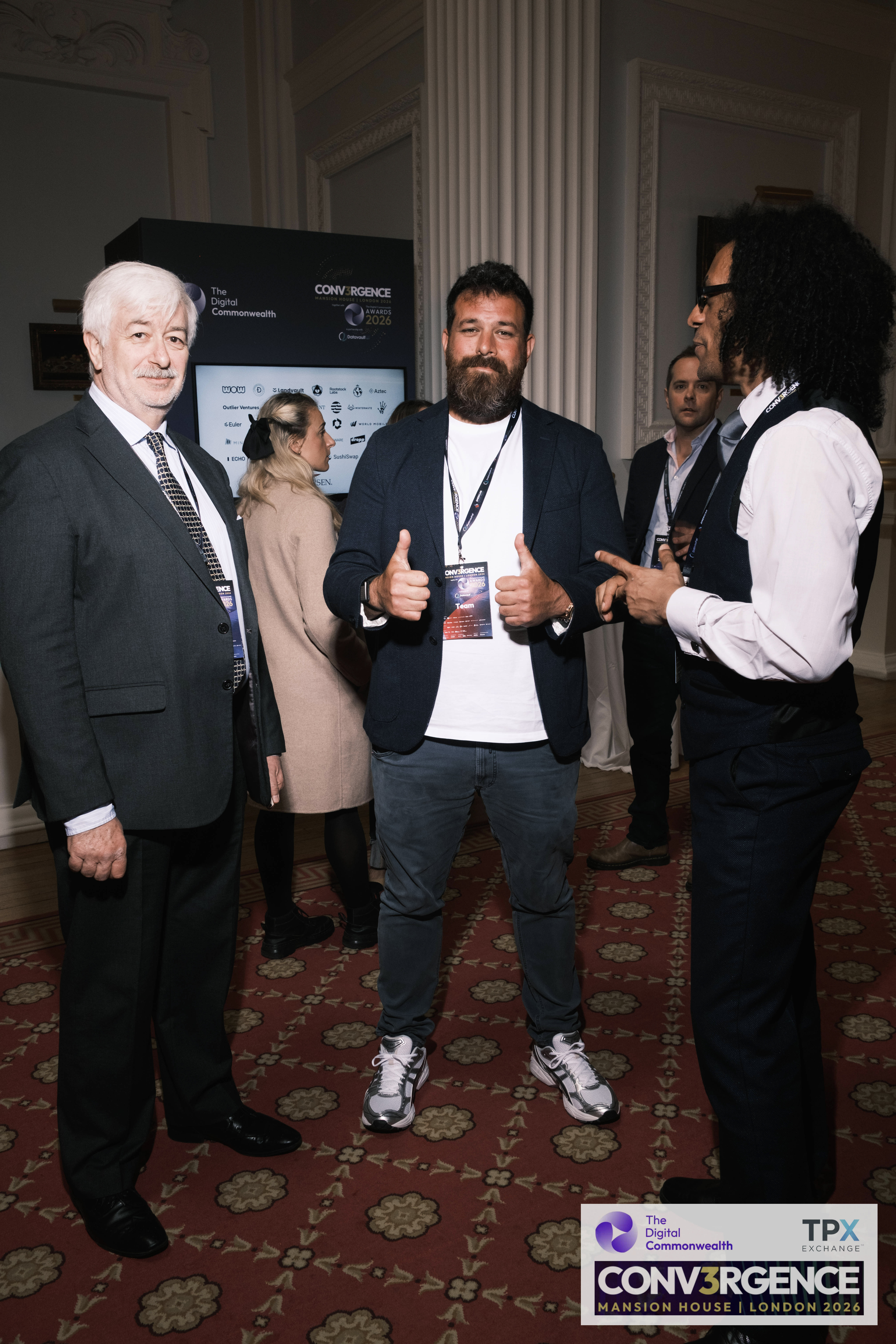 JB Webb-Benjamin in conversation with two other delegates at the Conv3rgence awards reception, Mansion House, in front of a screen of sponsor logos.