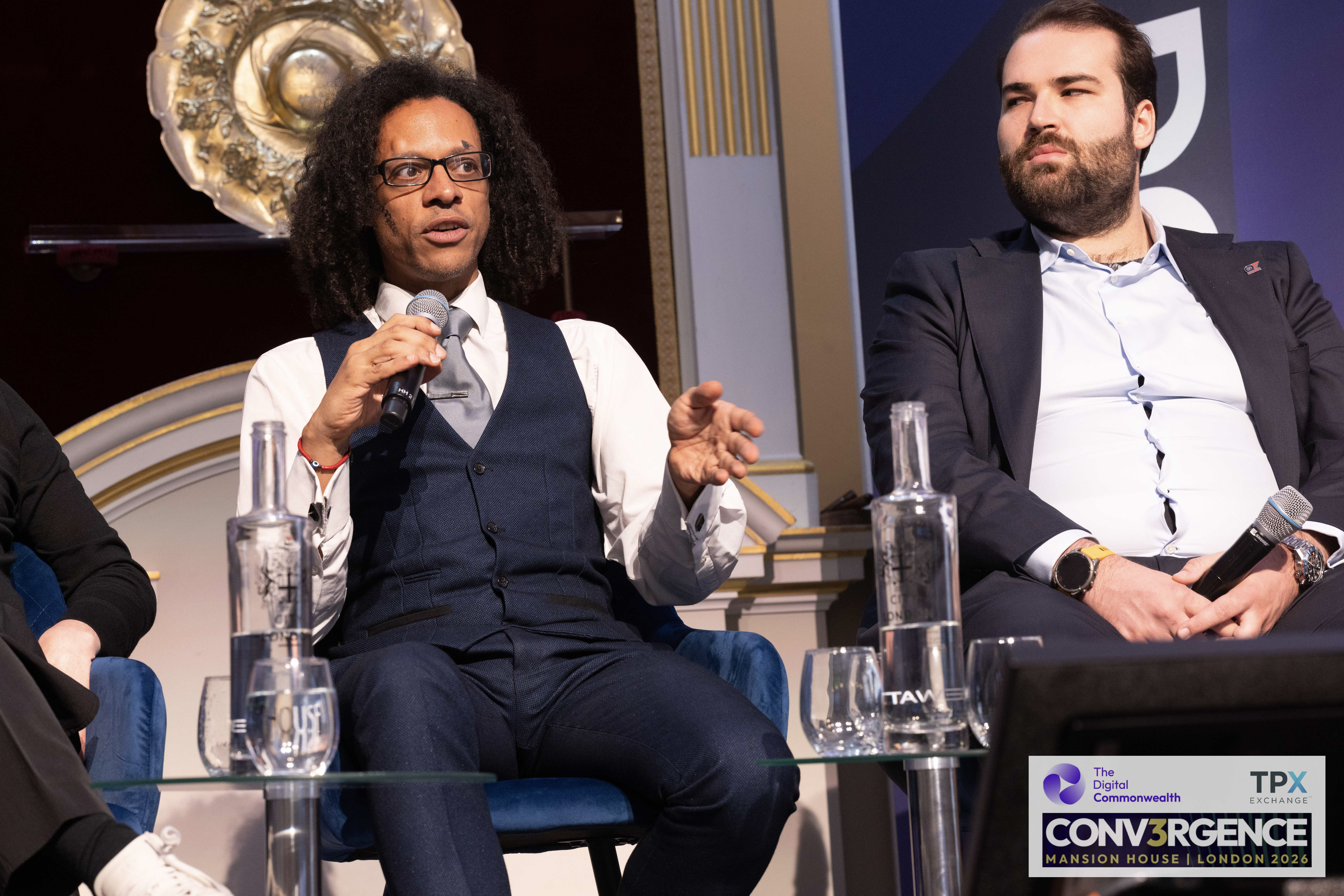 JB Webb-Benjamin speaking into a microphone on the international regulation panel at Conv3rgence, Mansion House, with a fellow panellist seated alongside.