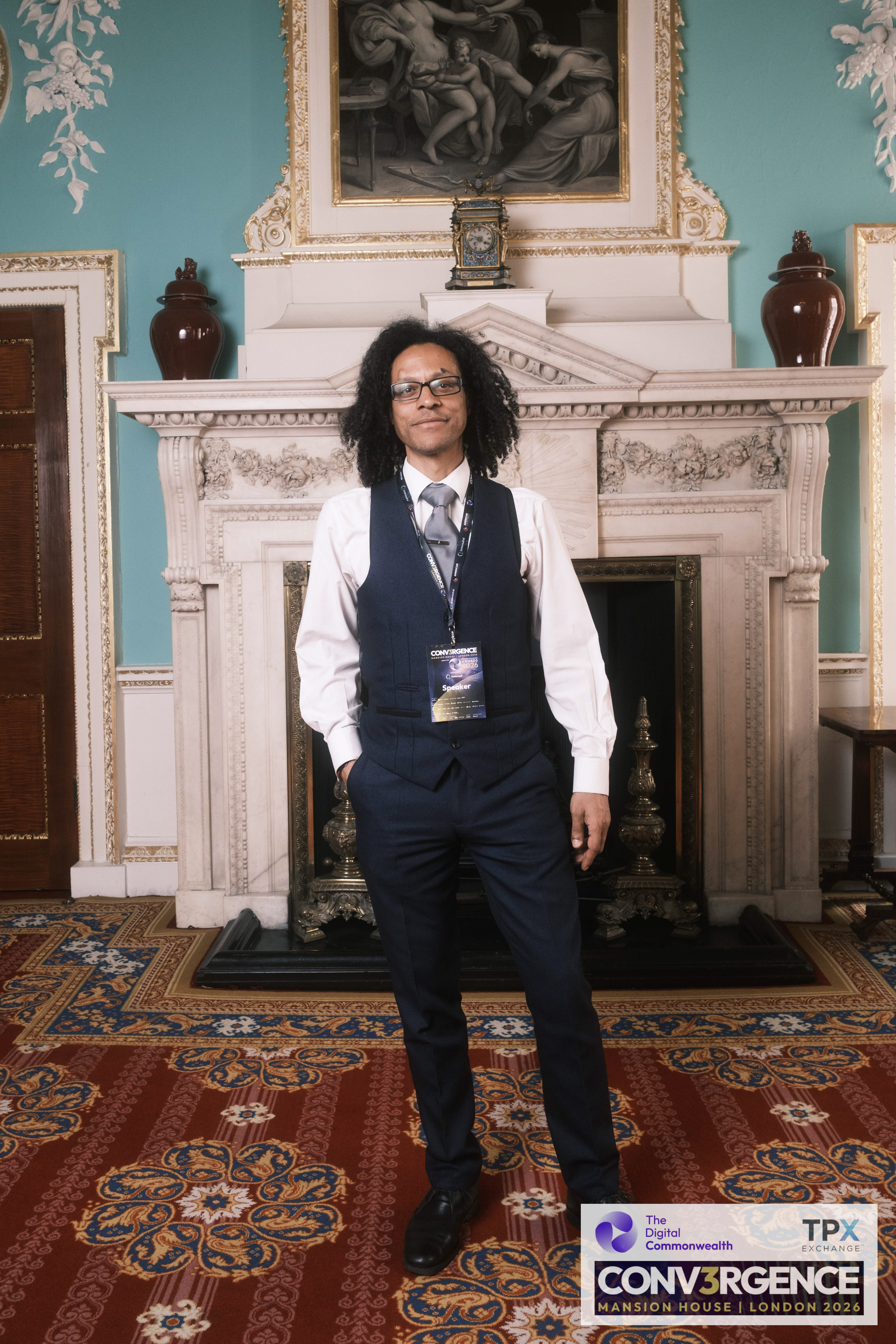 Full-length portrait of JB Webb-Benjamin standing by the marble fireplace in the Mansion House state rooms, wearing a Conv3rgence Speaker lanyard.