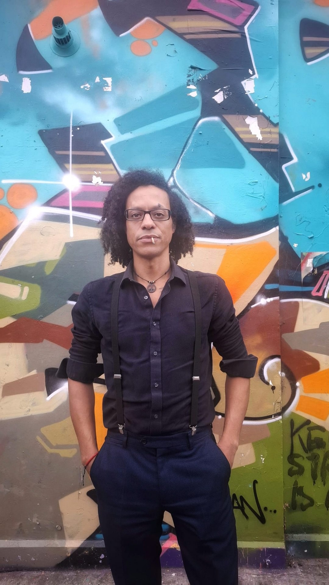 JB Webb-Benjamin in a black shirt and braces, glasses and shoulder-length curly hair, hands in pockets, standing in front of a large, brightly coloured graffiti wall.