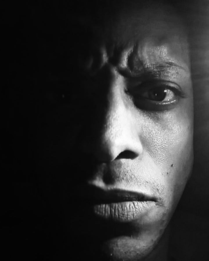 Dramatic black-and-white close-up portrait of JB Webb-Benjamin, half his face lit and the rest in deep shadow.