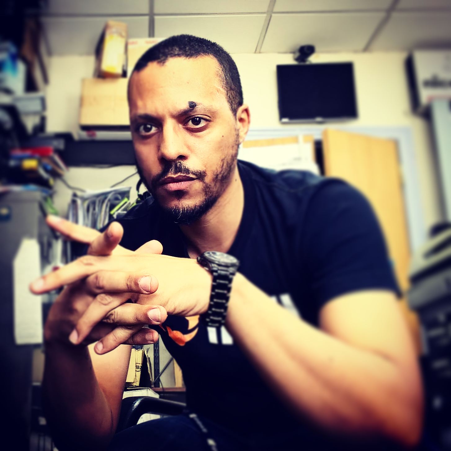 Candid portrait of JB Webb-Benjamin seated at a cluttered electronics workbench, hands clasped, wearing a black T-shirt and watch.