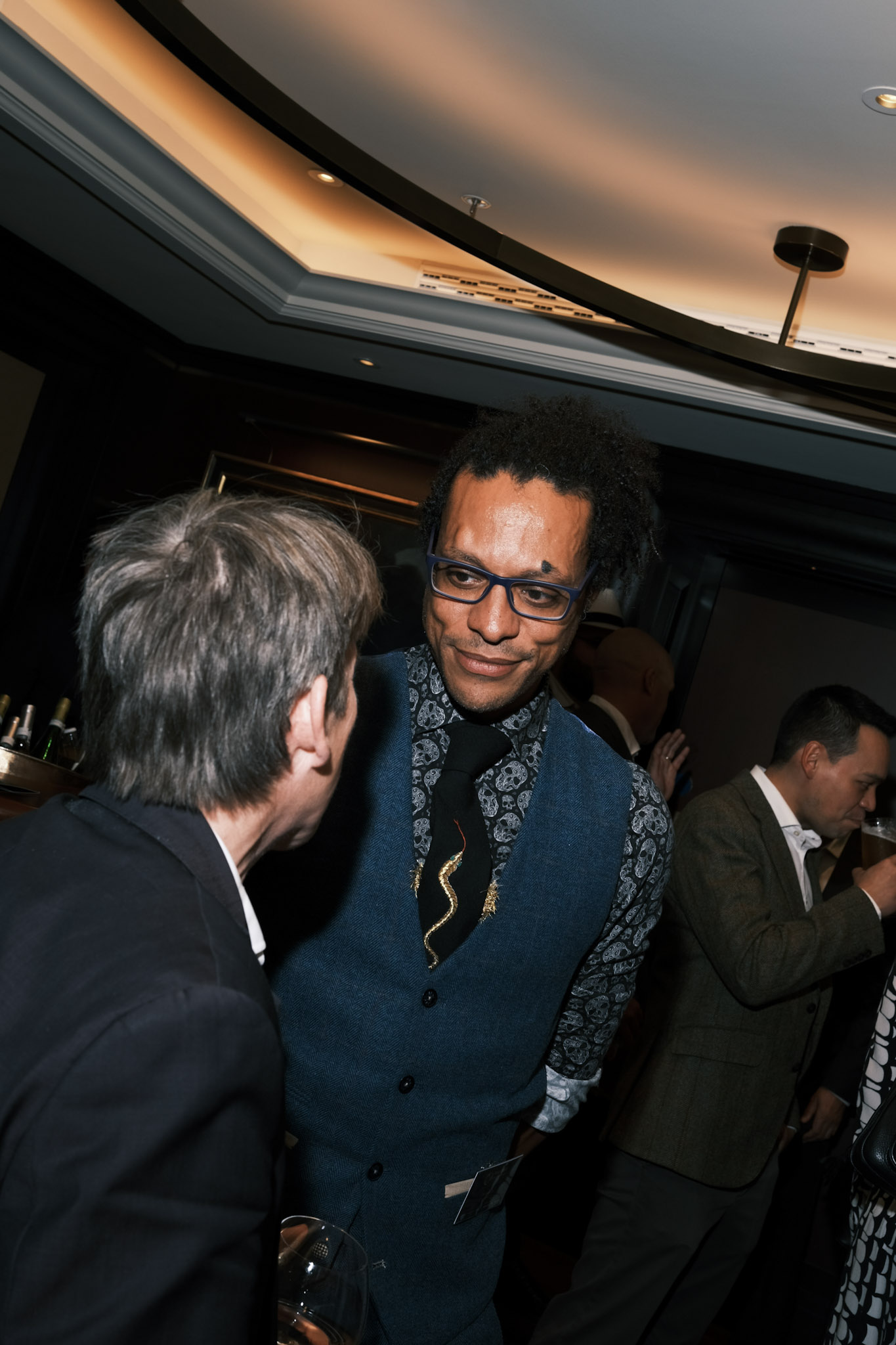 JB Webb-Benjamin smiling slightly as he listens to a guest seen from behind, with other guests and the bar in the background.