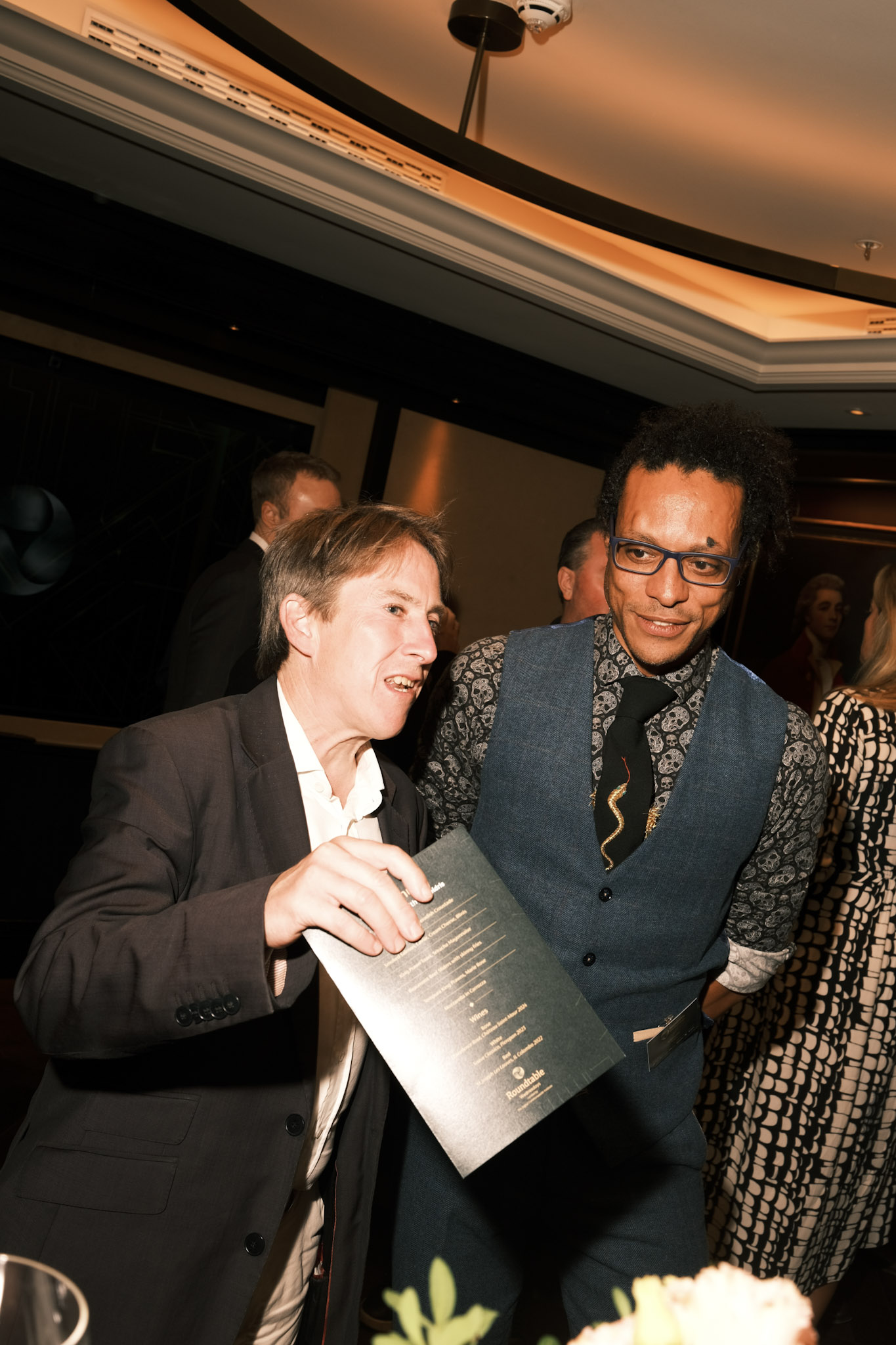 JB Webb-Benjamin leaning in beside a guest who is holding up the evening's printed Roundtable menu.