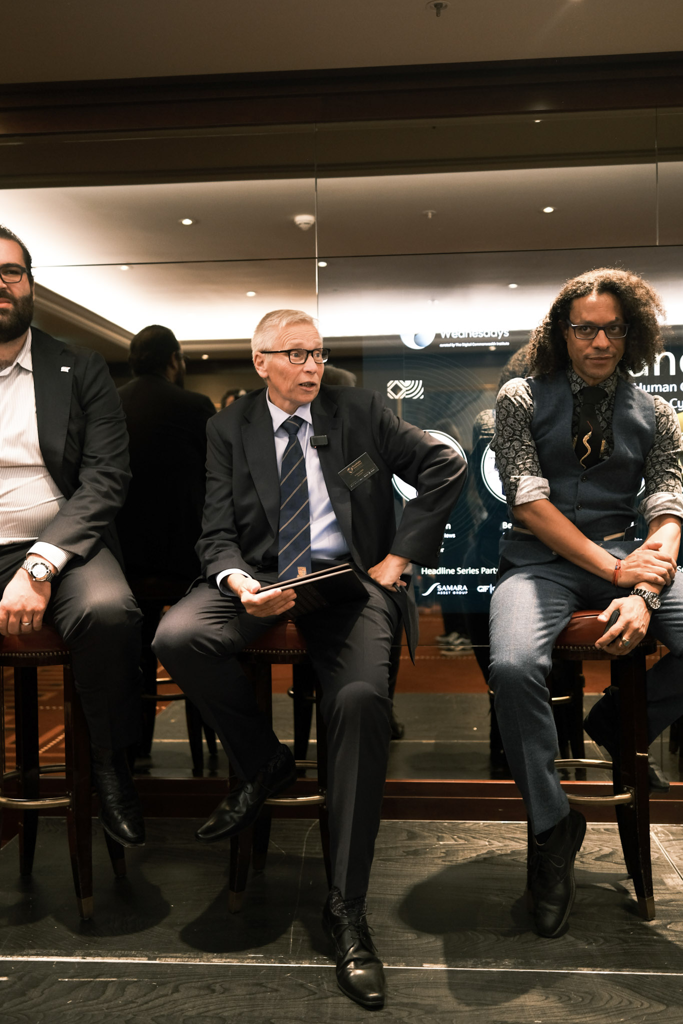 JB Webb-Benjamin seated on a bar stool at the right of a panel line-up, arms folded over one knee, looking at the camera, with two fellow panellists beside him and the Roundtable Wednesdays screen behind.