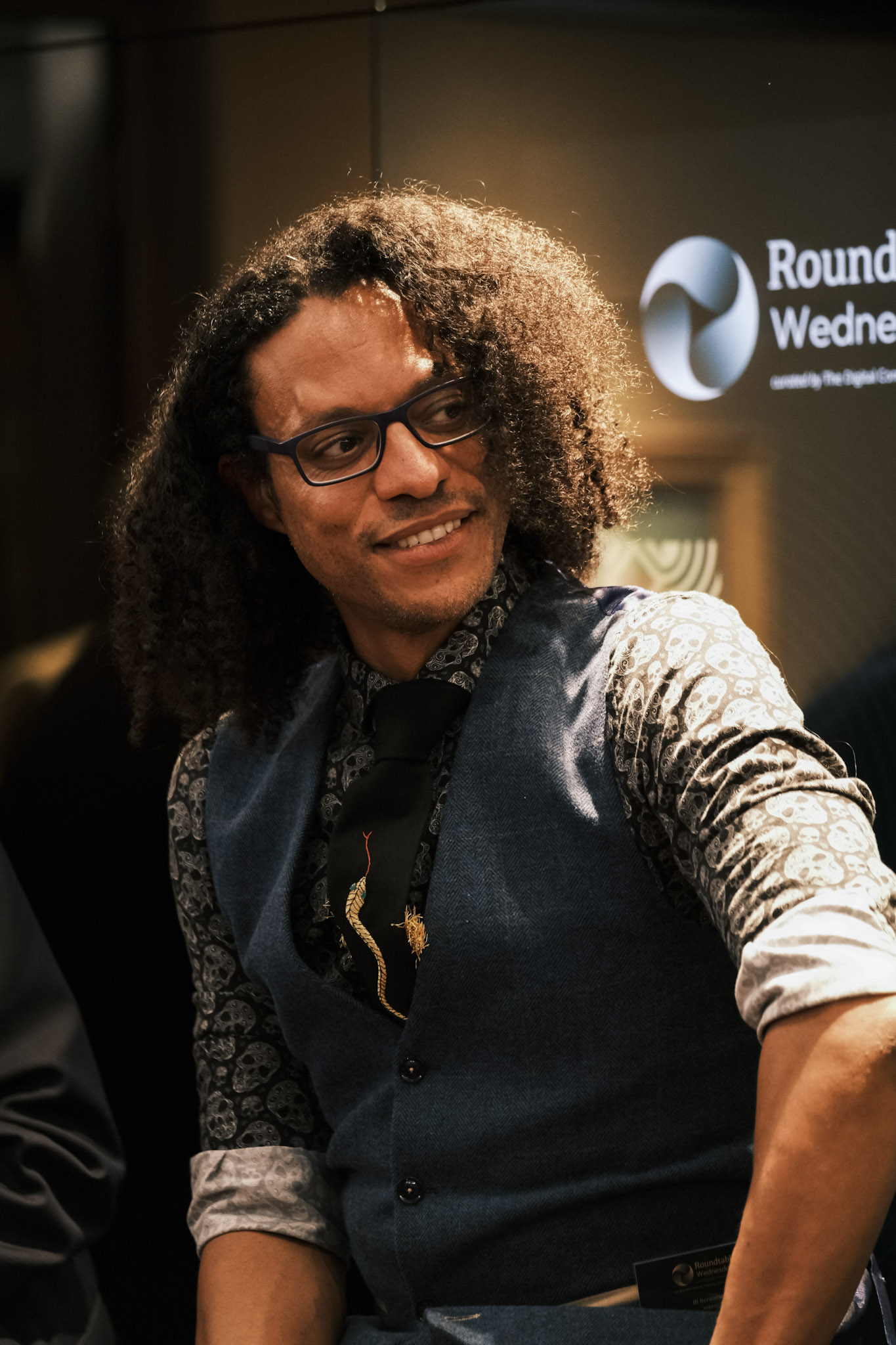 JB Webb-Benjamin smiling and glancing to one side, in glasses, a blue waistcoat, dark skull-print shirt and an embroidered tie, with the Roundtable Wednesdays logo behind.