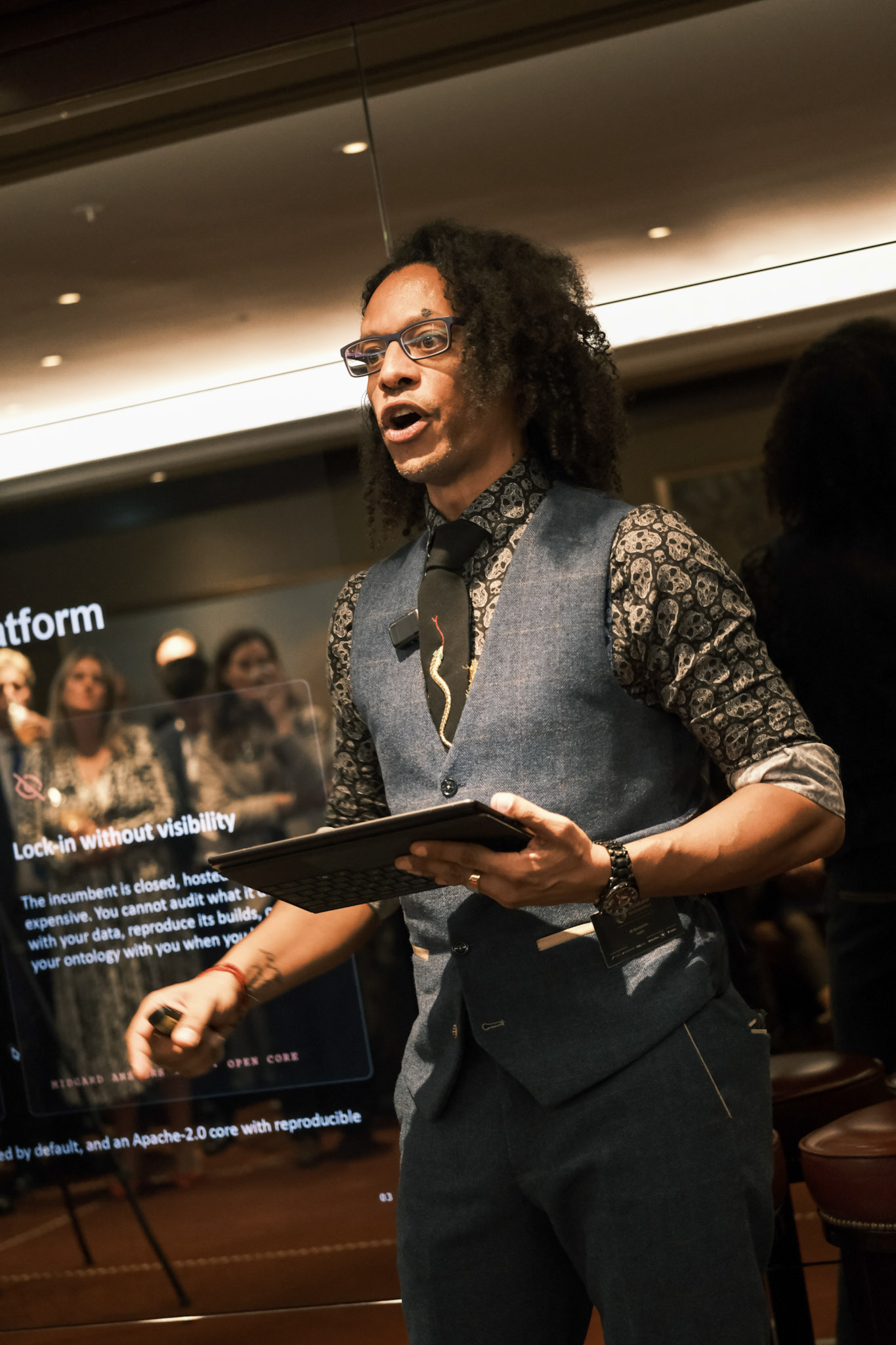 JB Webb-Benjamin in a blue tweed waistcoat and skull-print shirt, holding a tablet keyboard and a clicker, presenting in front of a large screen showing a Midgard slide headed 'Lock-in without visibility'.