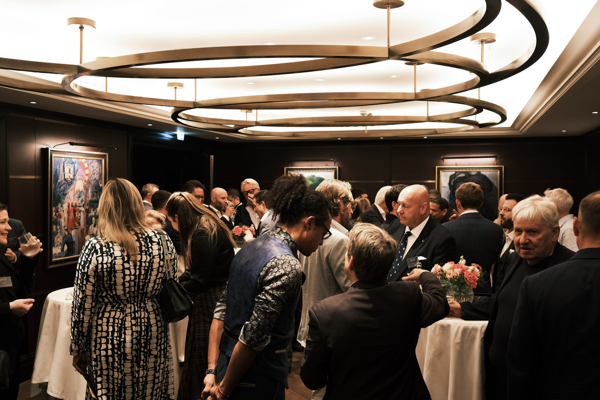 A busy reception room with large ring-shaped ceiling lights, guests gathered around white-clothed tables, and JB Webb-Benjamin in the middle of the crowd, bowed in conversation.