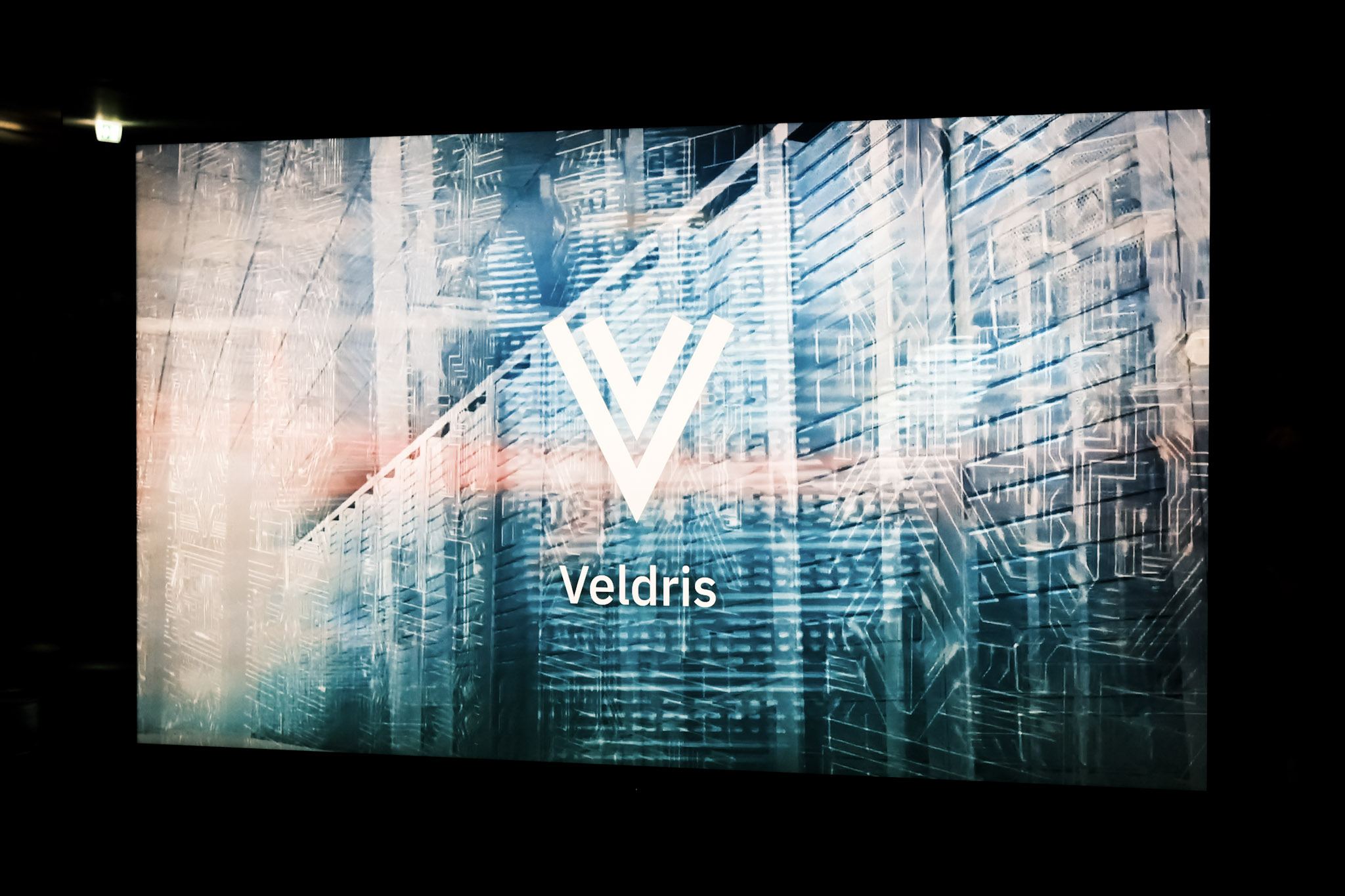 A large screen in a dark room showing the white Veldris V logo and wordmark over a blue-toned image of server racks.