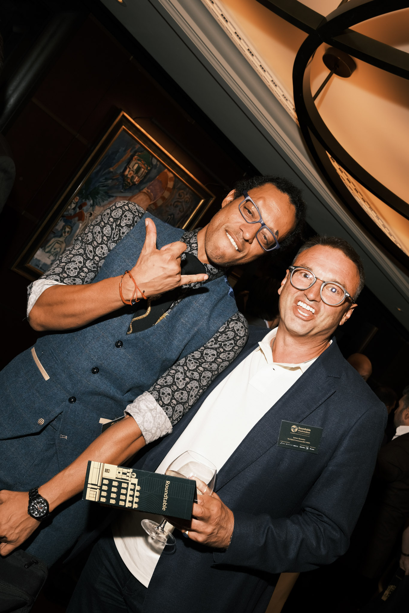 JB Webb-Benjamin leaning in and making a shaka hand sign beside a grinning fellow guest holding a Roundtable programme.